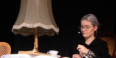 Powiększ grafikę: Kobieta siedzi przy biurku, na którym leży książka, filiżanka i dzwonek.