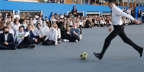 Powiększ grafikę: Pan Jarosław Kovac kopie piłkę nożną. Uczniowie siedzą na podłodze.